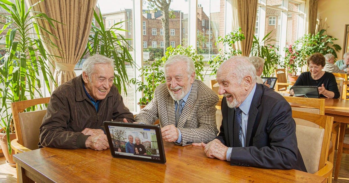 Three older men laughing together while watching a video call on a large-screen tablet in a bright care home lounge