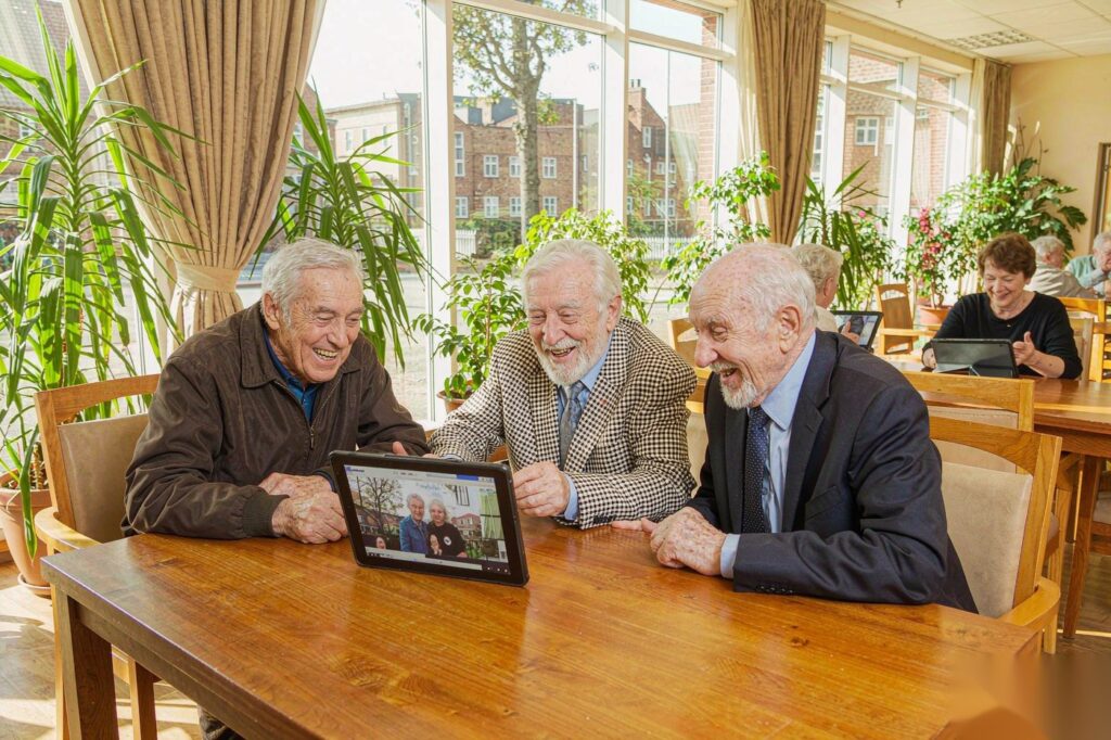 Three older men smiling at a large-screen tablet showing a family photo album in a sunny communal lounge
